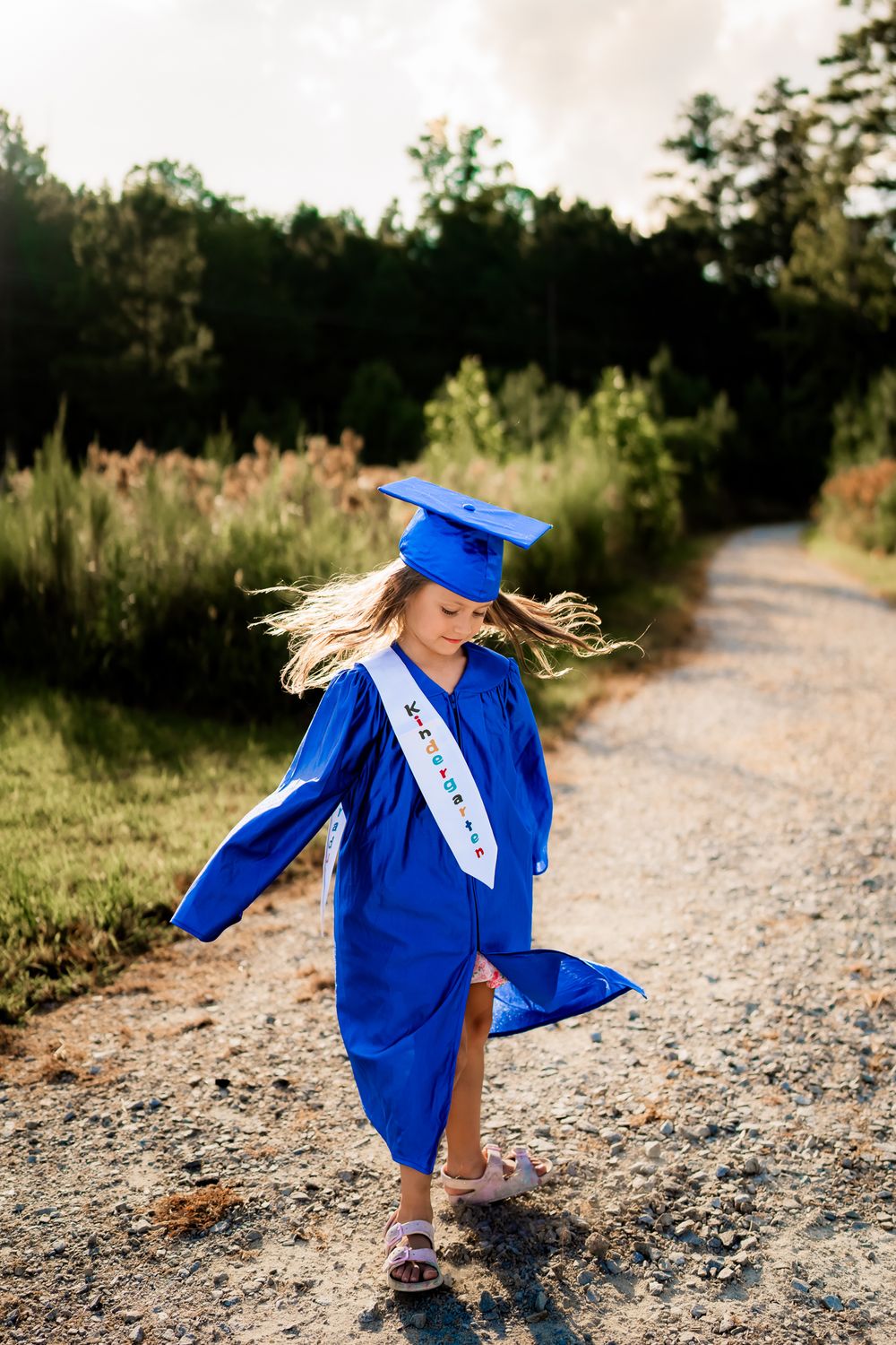 Graduate in royal blue cap and gown twirls playfully on a gravel path with trees and wildflowers in background.