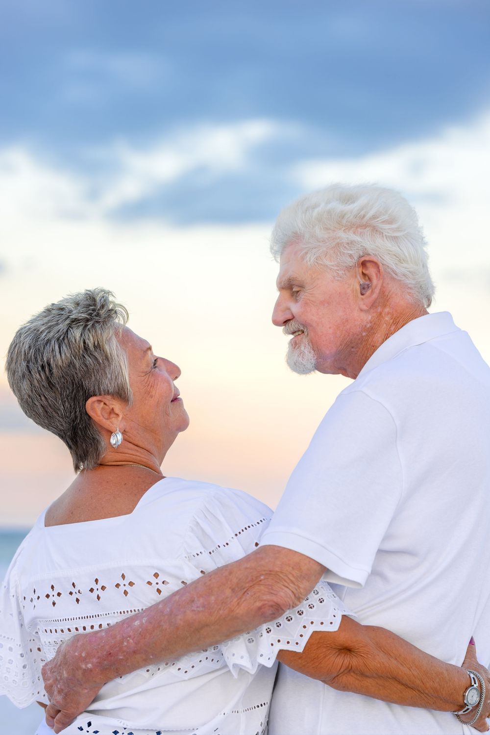 Two seniors in white outfits share affectionate moments on a beach at sunset with soft clouds in the background.