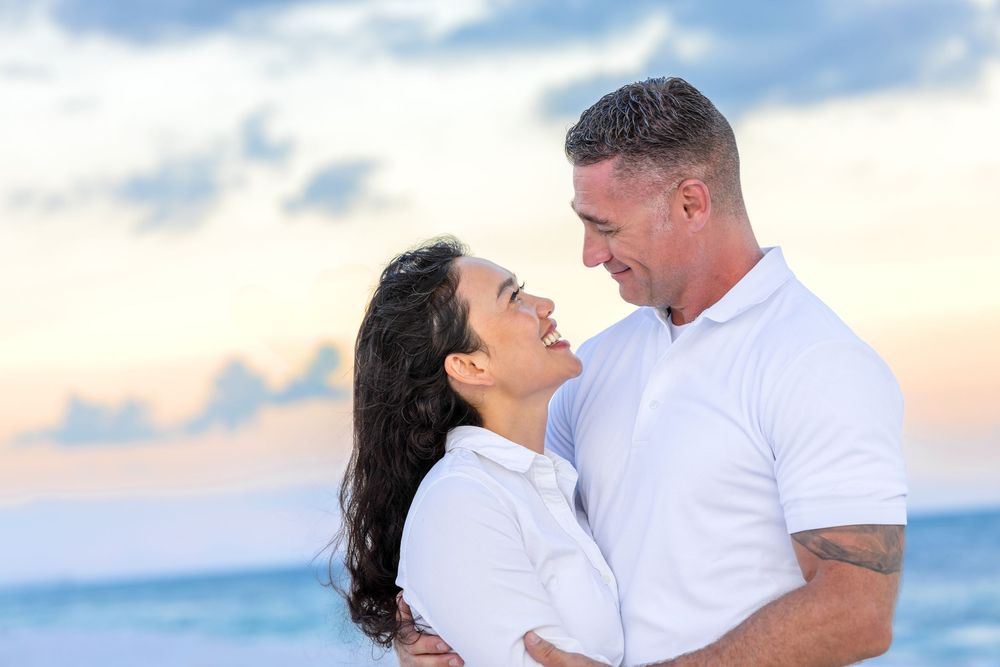 Loving couple in white attire embraces against scenic beachside background during dreamy sunset photoshoot.