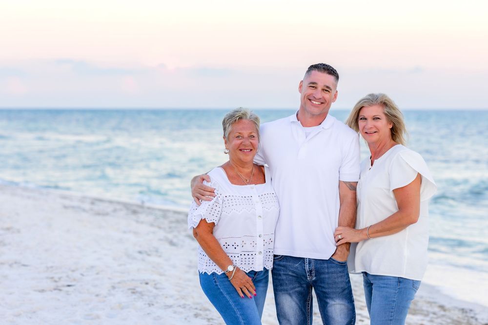 Family enjoying a casual beach photoshoot in white and denim outfits at sunset along the ocean shore.