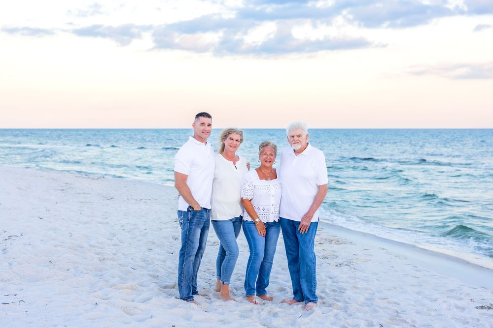 Group wearing white shirts and blue jeans pose together on a sunny beach with ocean waves in background.