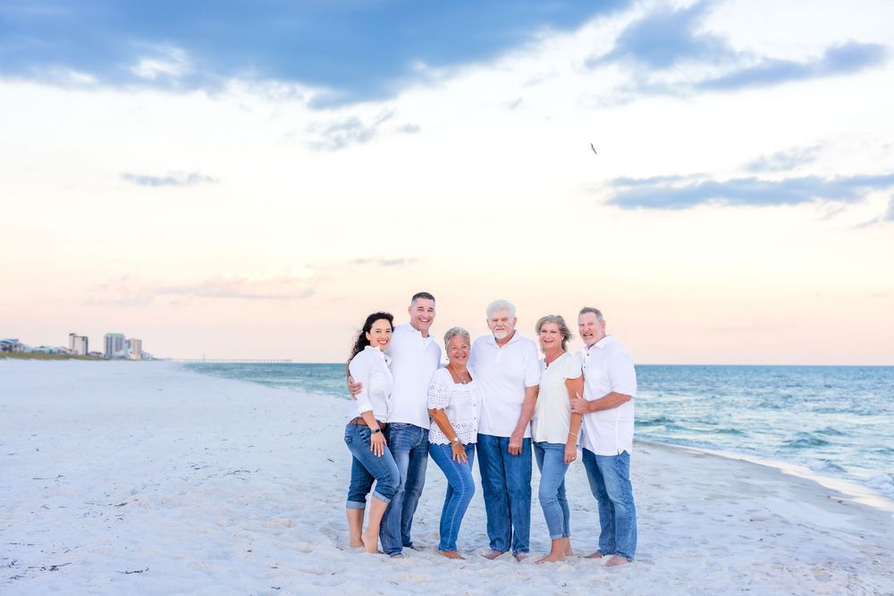 Family gathering at sunset on white sandy beach with coastal buildings visible in distance.
