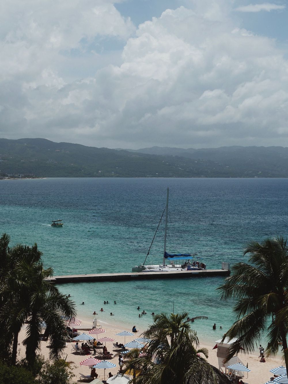 Stunning panoramic view of a Caribbean beach with turquoise waters, palm trees, and boats against mountains in Jamaica.