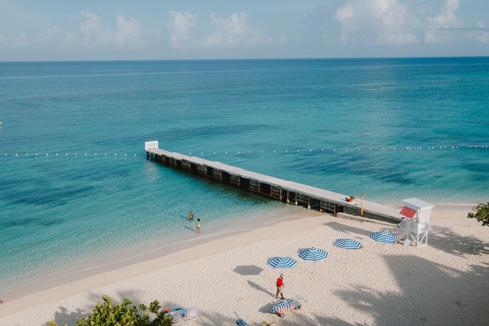 Row of pristine beach umbrellas line the shoreline of a turquoise Caribbean ocean with a wooden pier stretching seaward.