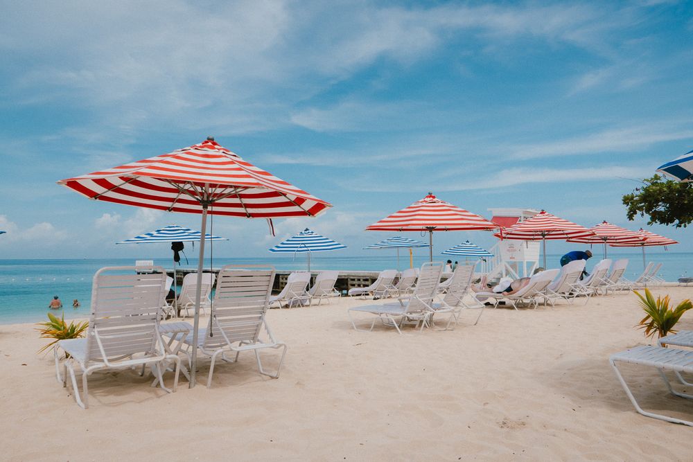 Red and white striped beach umbrellas line the sandy shoreline with blue sky and ocean views in a panoramic photo.