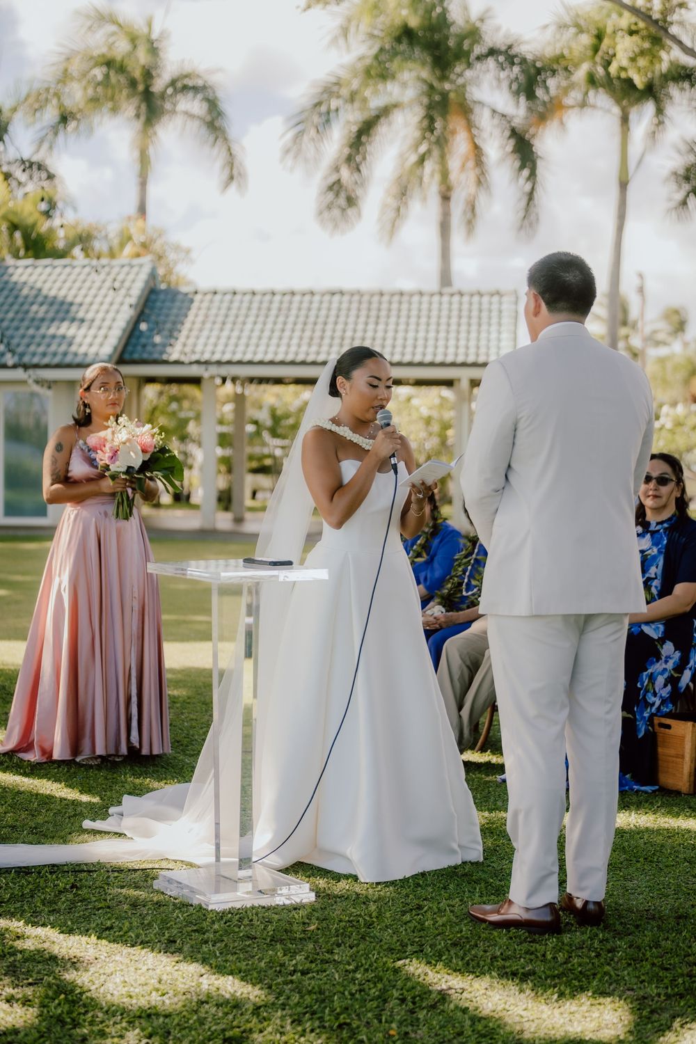 Wedding guests gather on a sunny lawn with palm trees and a green-roofed building in the background.