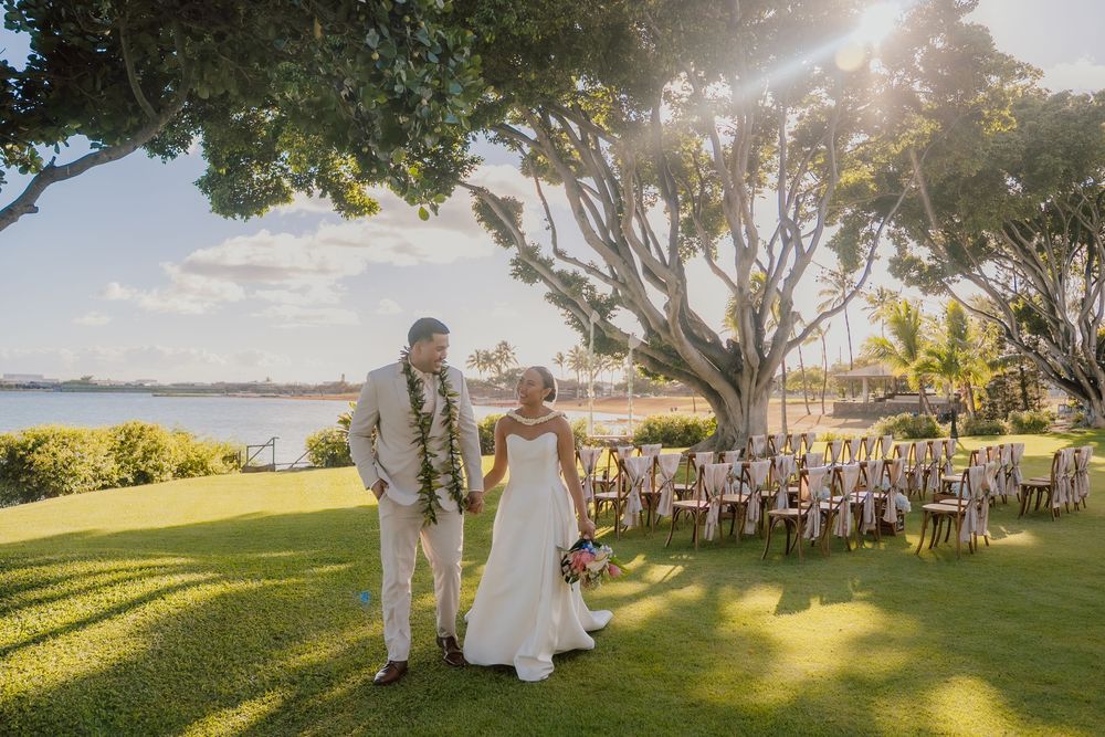 Outdoor wedding ceremony setup with guests seated on a grassy hillside overlooking the ocean at sunset.