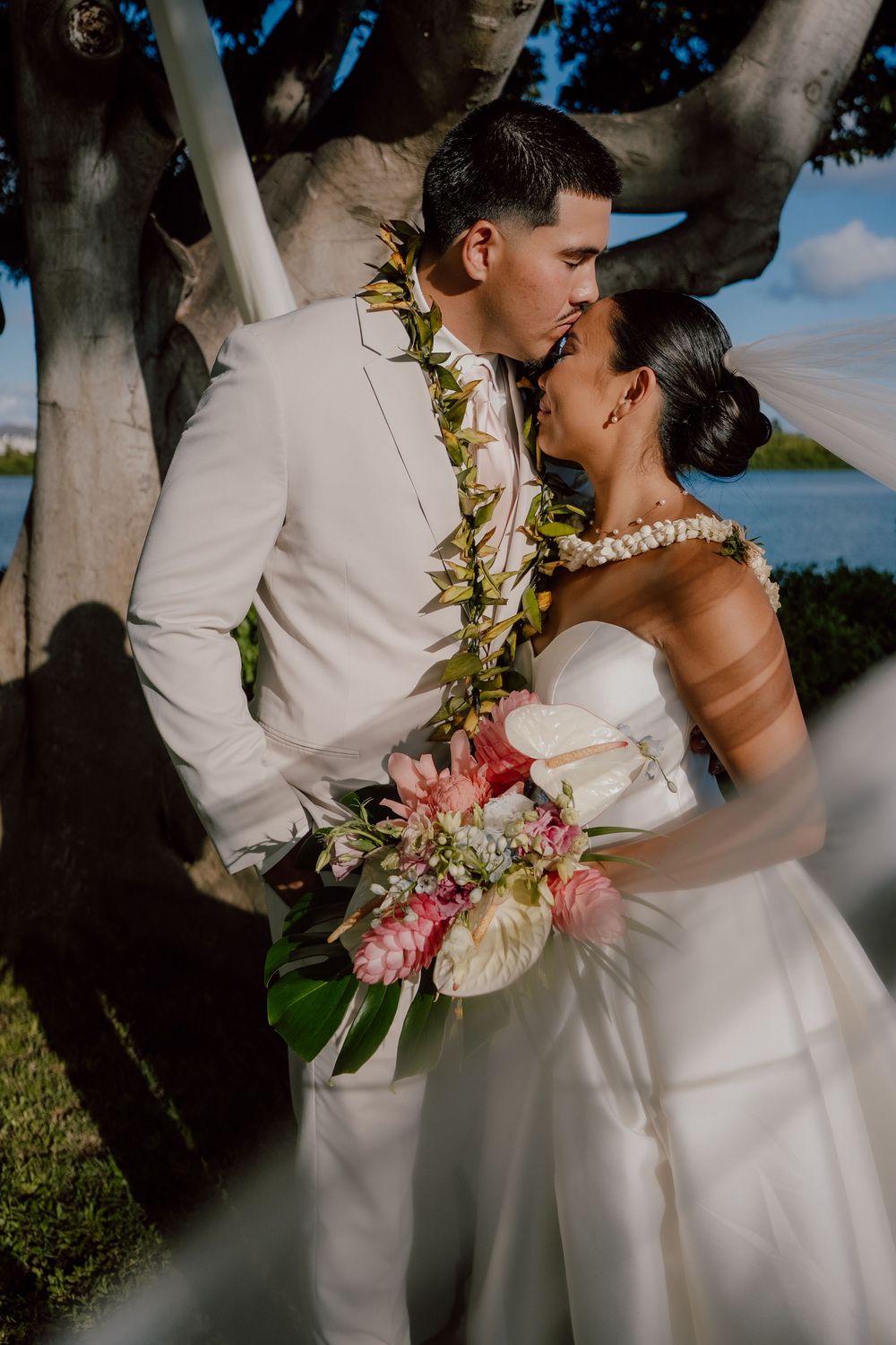 A romantic wedding couple embraces under trees during an outdoor ceremony with tropical flowers and white attire.
