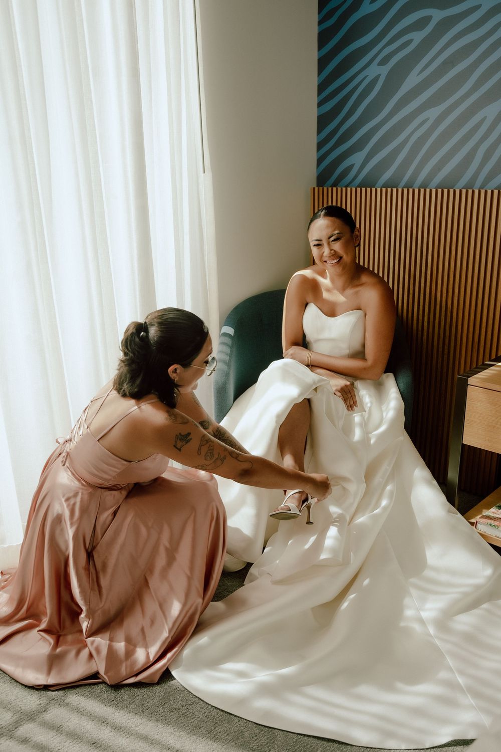 A bridesmaid in a pink dress helps adjust the flowing white wedding gown as they prepare for the ceremony.
