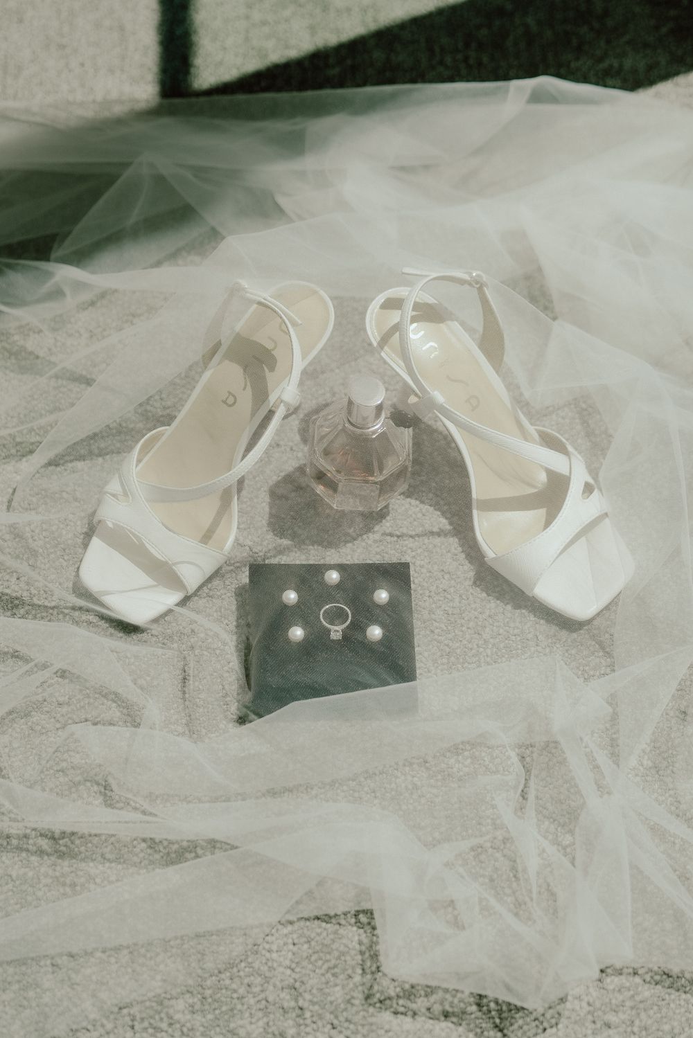 White wedding shoes, perfume bottle and rings arranged on delicate tulle fabric in elegant bridal styling.