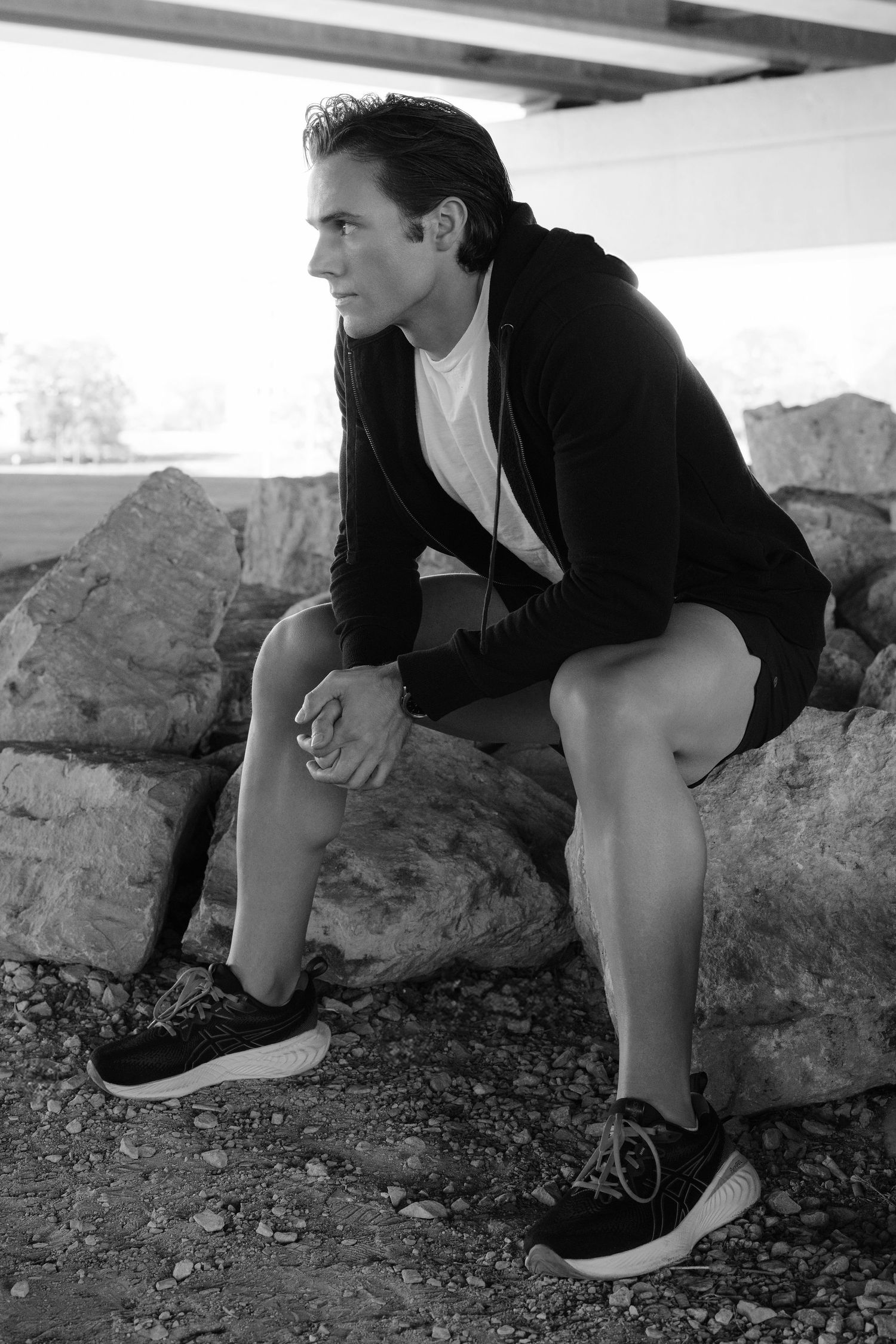 Athletic figure in hoodie sits contemplatively on rocks in dramatic black and white composition.