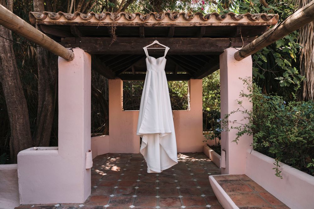 White wedding dress hanging in pink archway against natural outdoor backdrop.