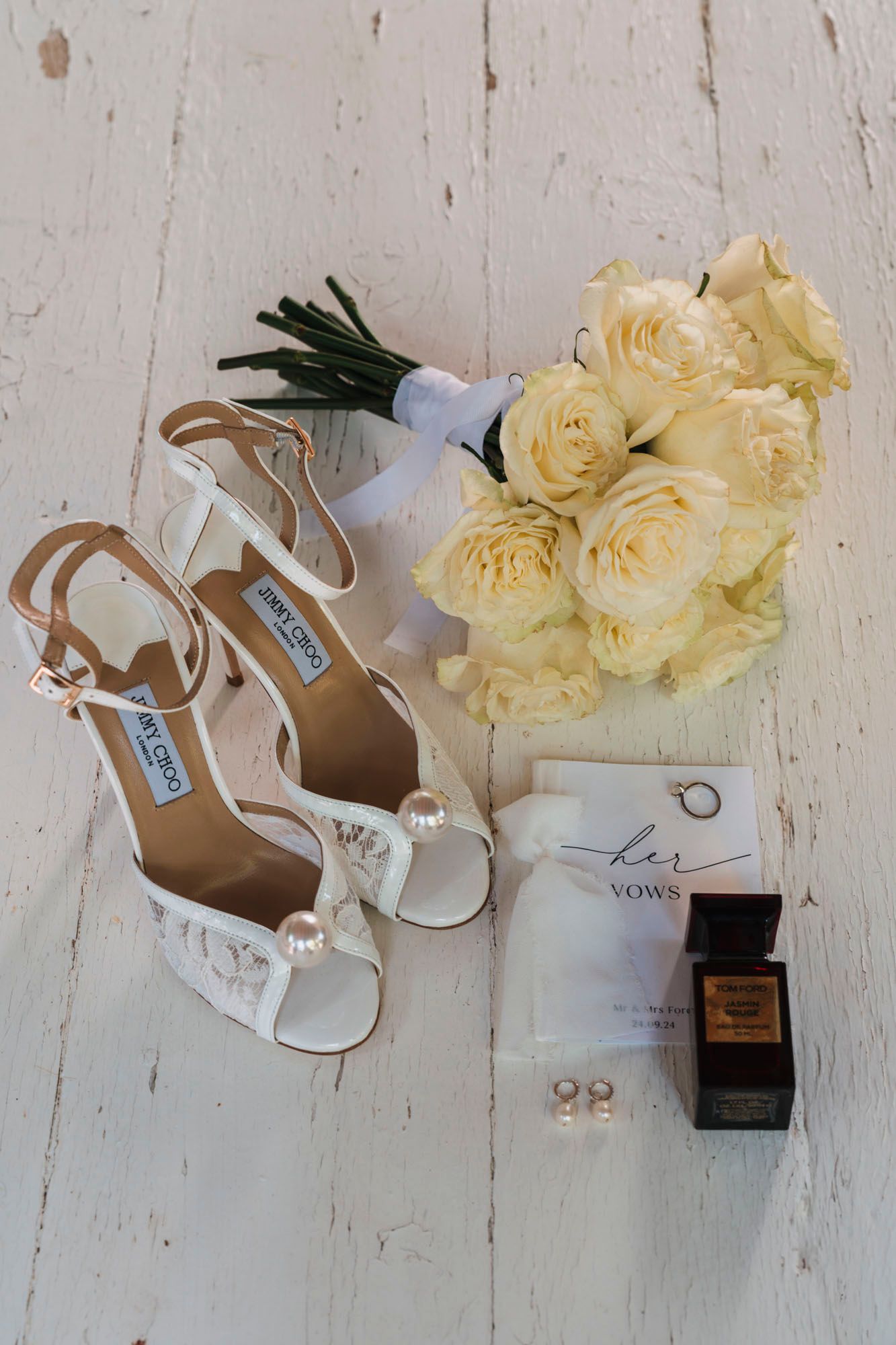 Wedding details including white bridal shoes, yellow roses, and perfume arranged on a white wooden surface.