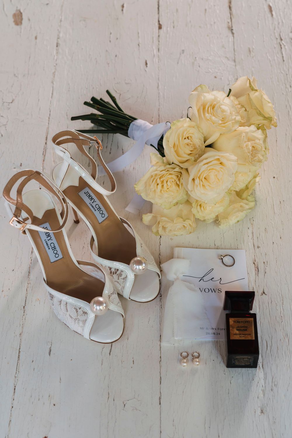 Wedding details including white bridal shoes, yellow roses, and perfume arranged on a white wooden surface.