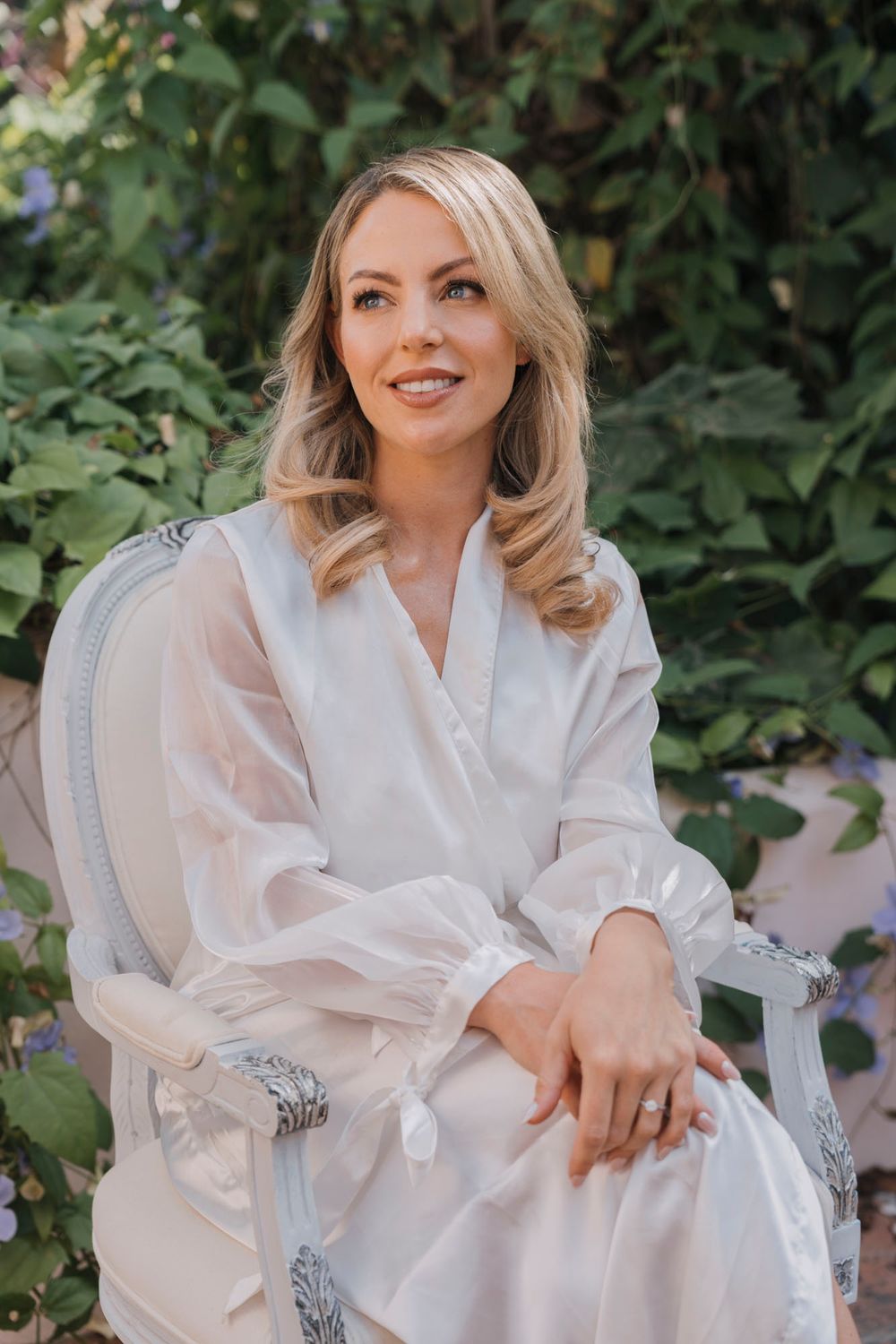 A woman in a white robe sits elegantly in a garden chair surrounded by lush greenery.