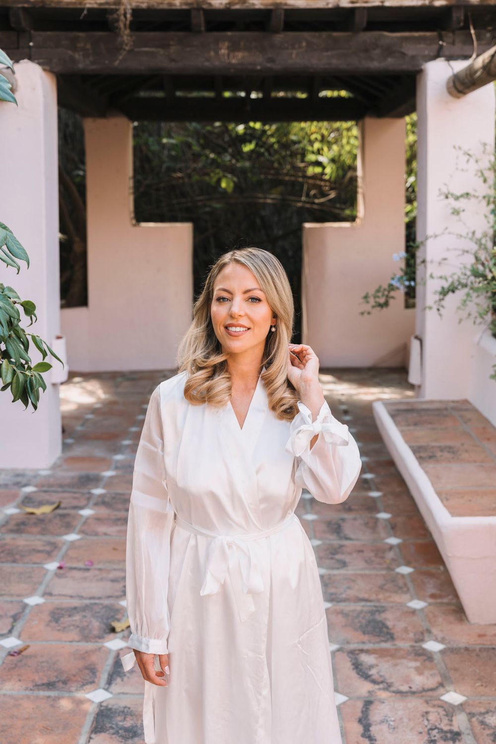 A woman in a white robe poses in a Mediterranean-style courtyard with stone pavement.