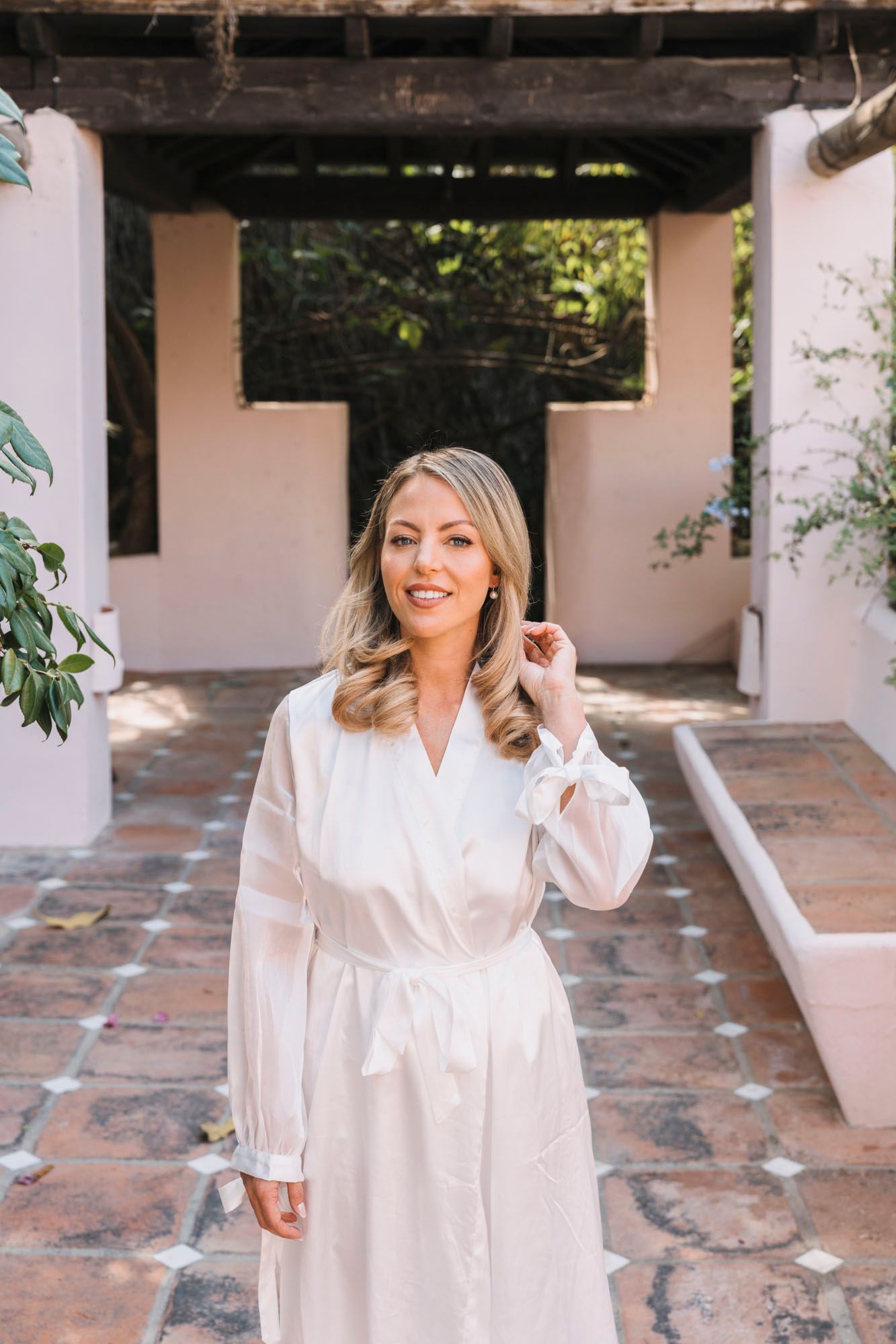 A woman in a white robe poses in a Mediterranean-style courtyard with stone pavement.