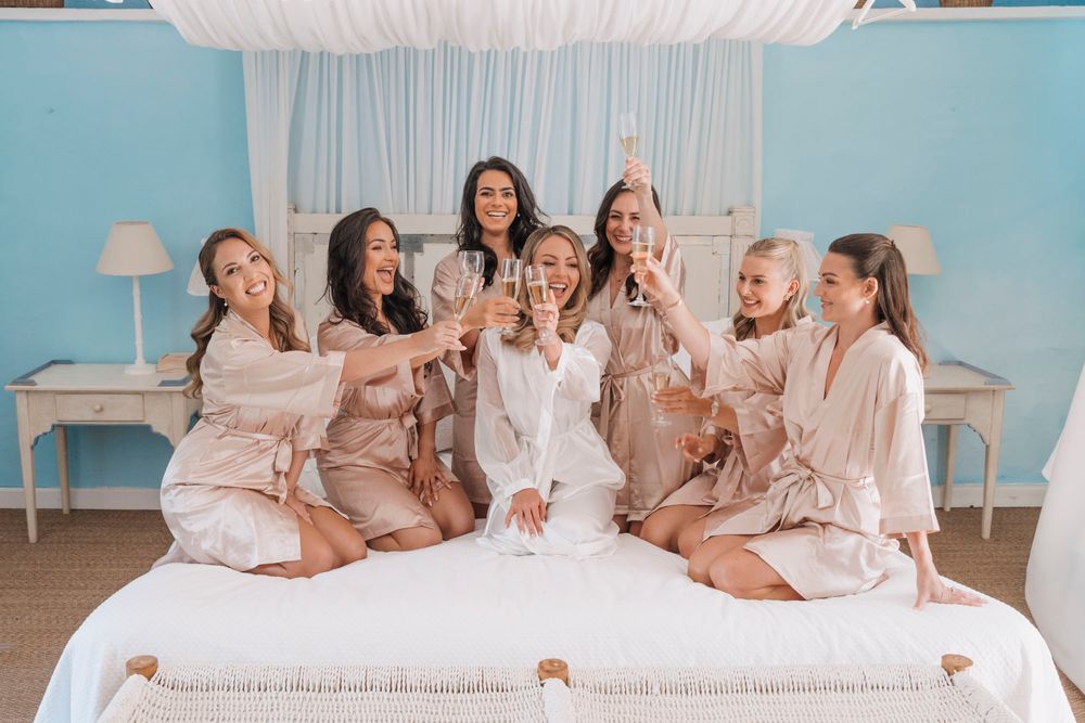 Wedding party in silk robes posing playfully on white bed during pre-ceremony preparations.