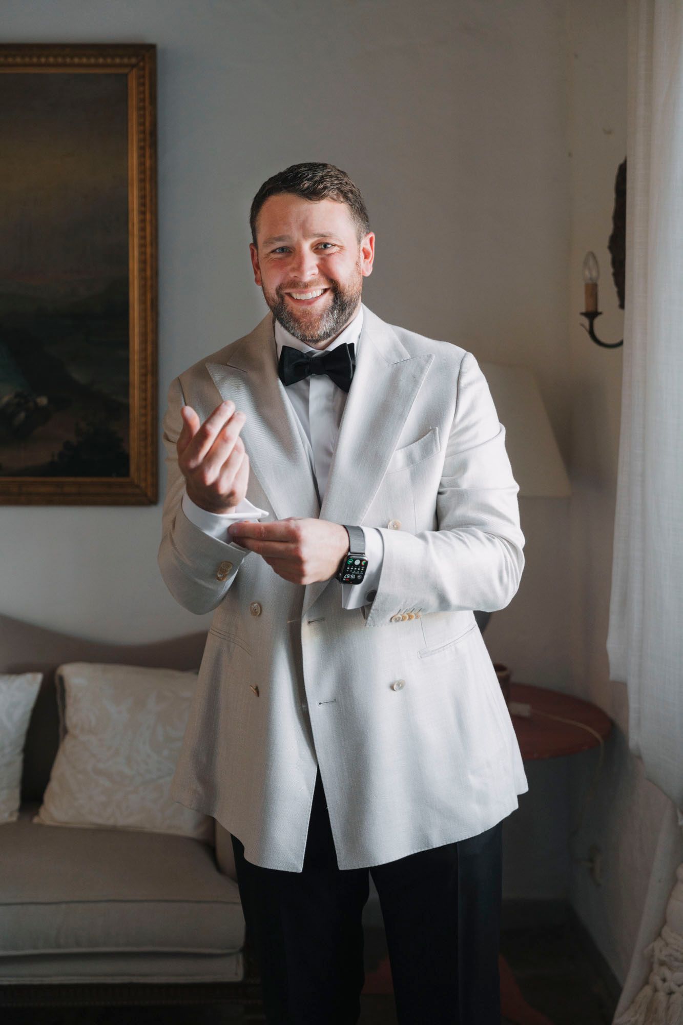 Person in white dinner jacket and bow tie smiling in formal interior setting.