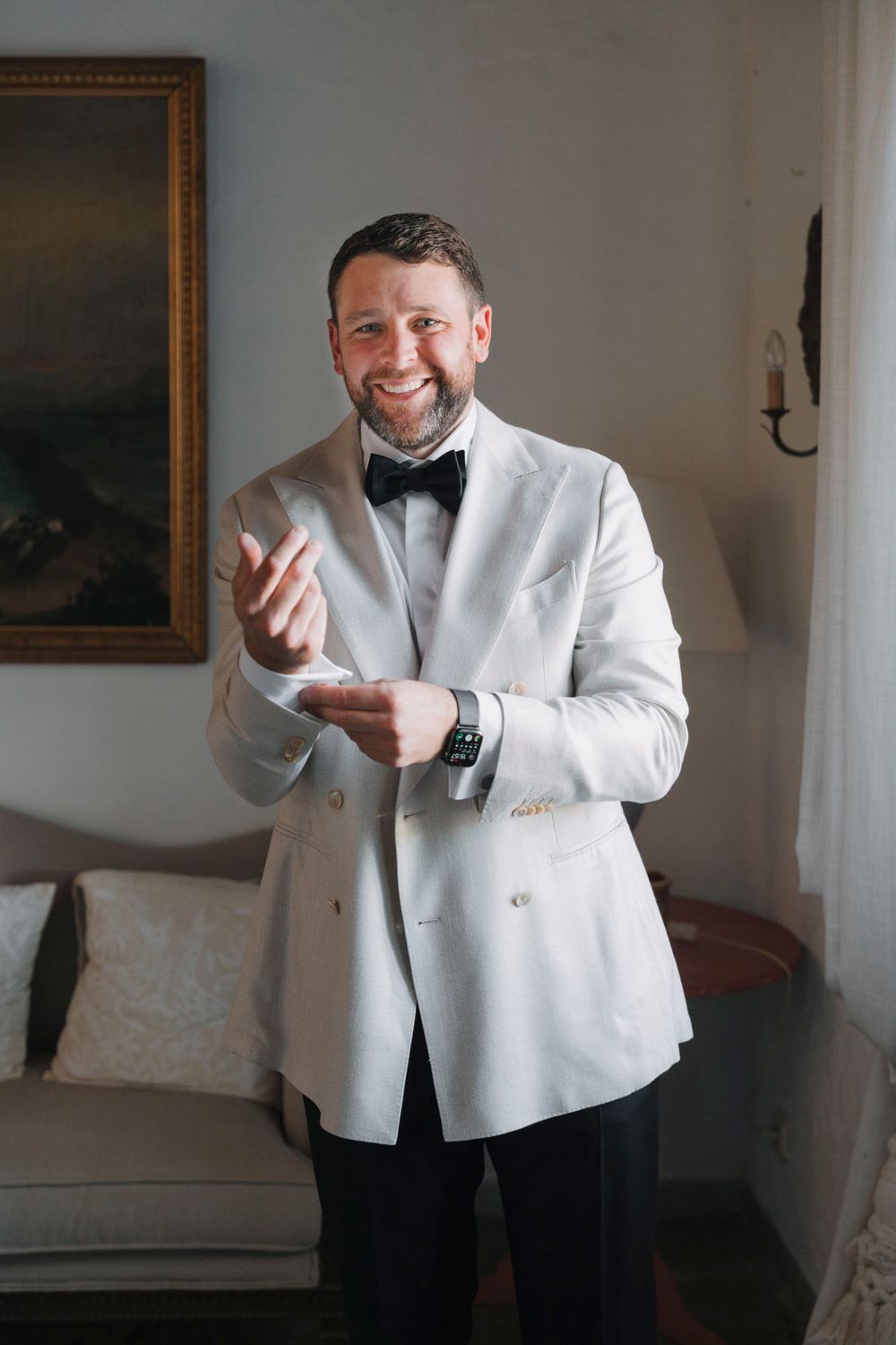 Person in white dinner jacket and bow tie smiling in formal interior setting.