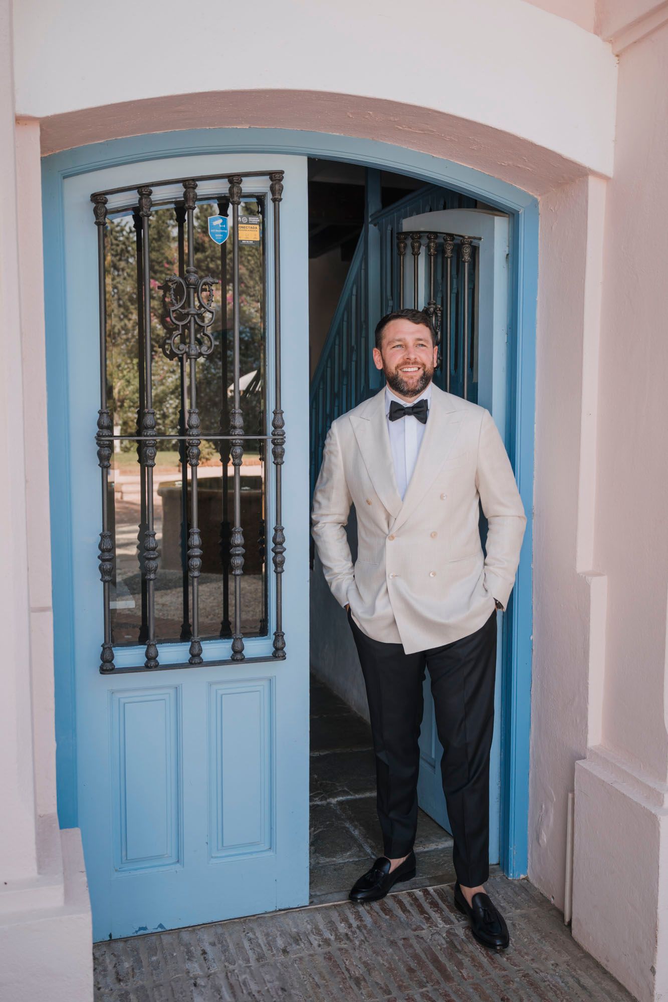 Groom in cream blazer and bowtie stands confidently in blue doorway arch at outdoor venue.