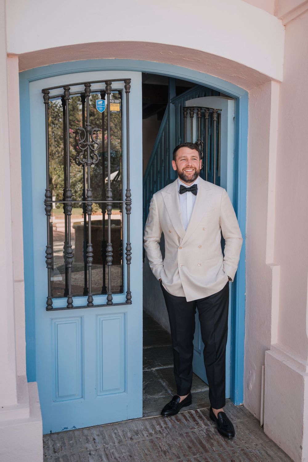 Groom in cream blazer and bowtie stands confidently in blue doorway arch at outdoor venue.