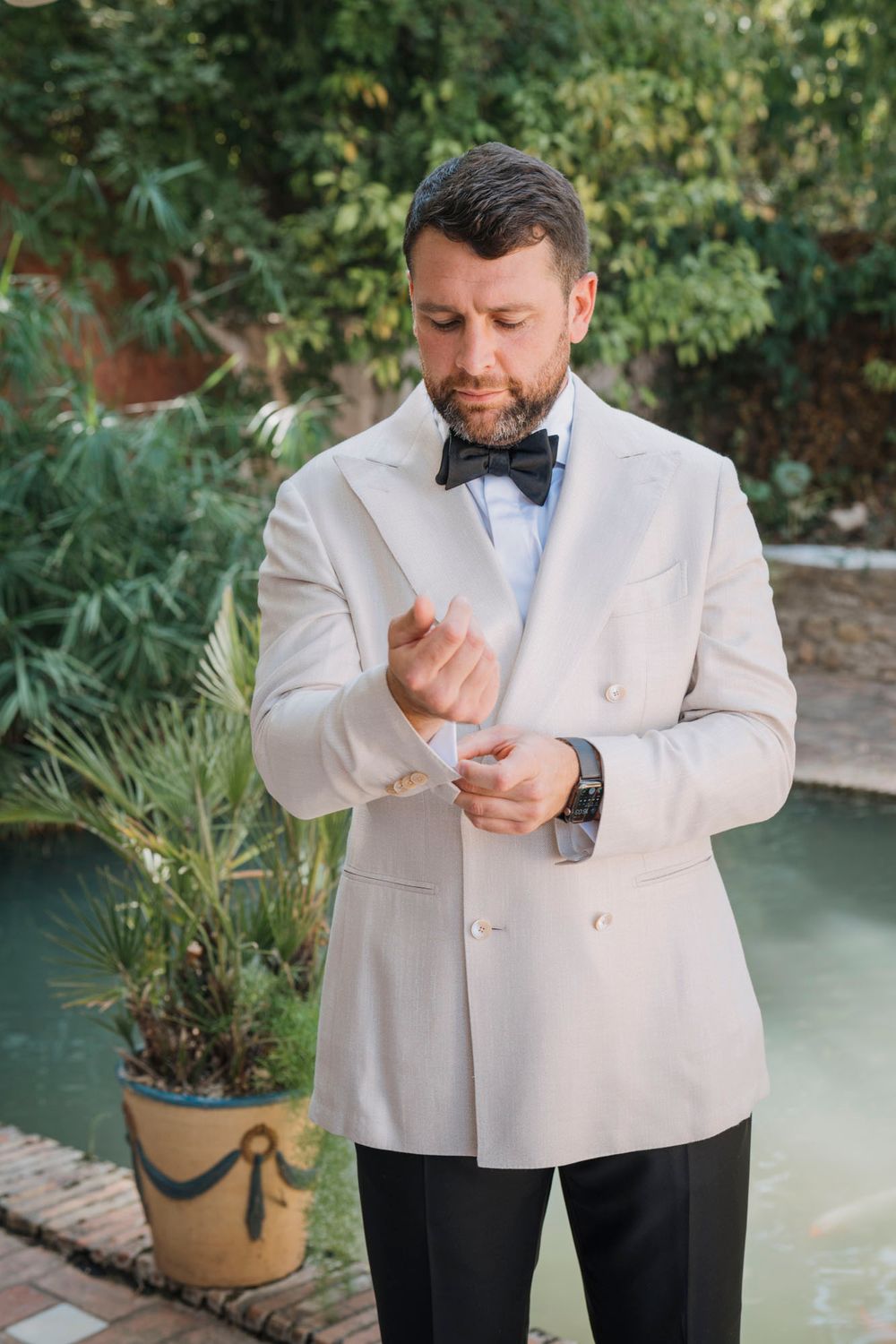 Groom adjusts cuffs of white double-breasted jacket while standing by garden setting.