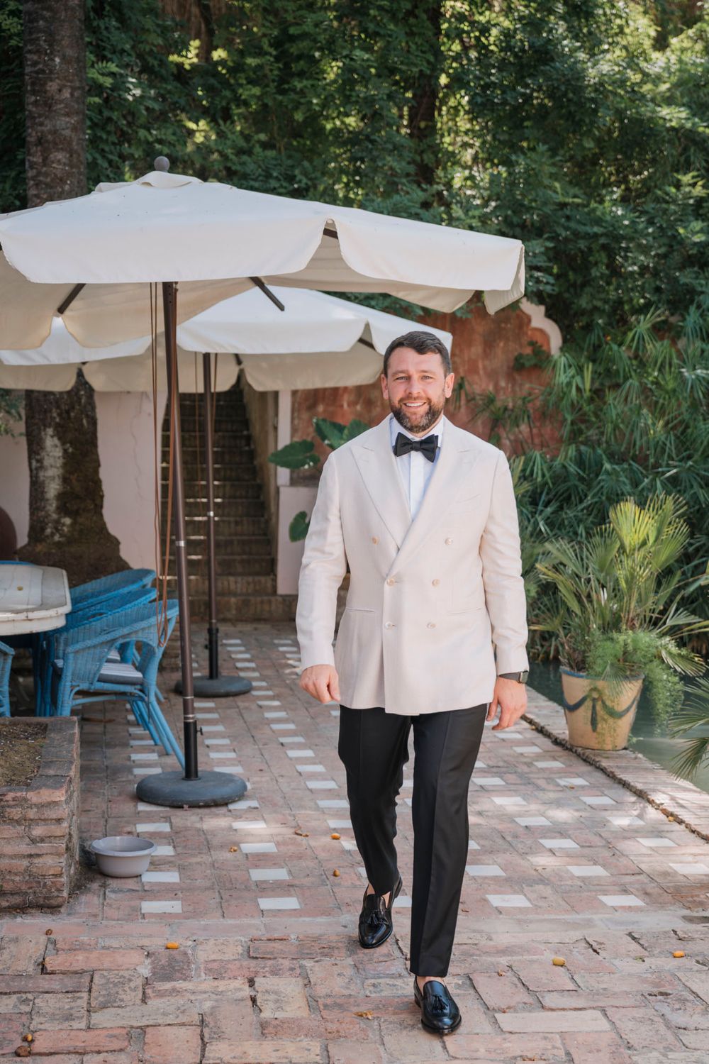 Elegantly dressed man in cream jacket walks along brick pathway under white umbrellas.