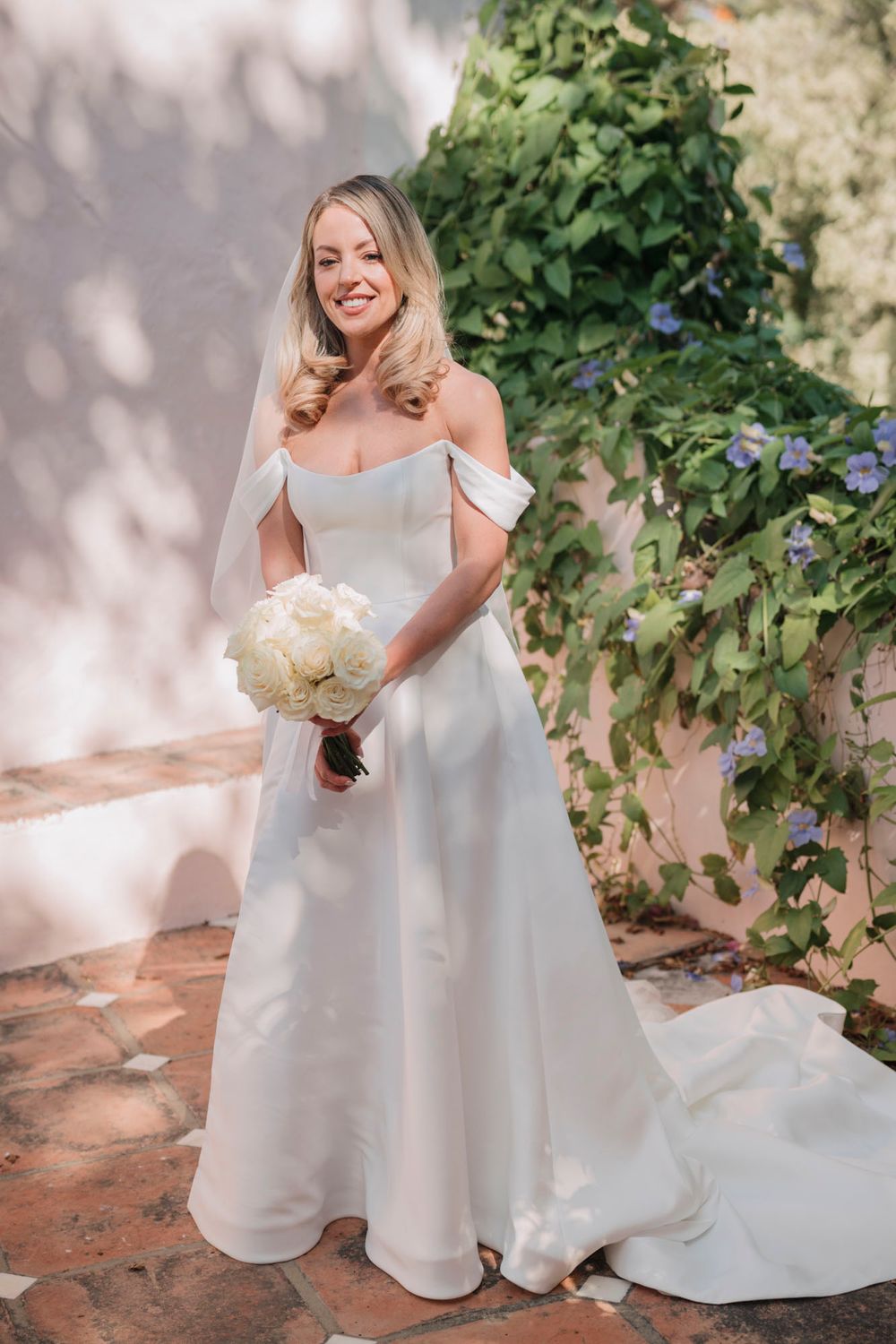Bride in classic off-shoulder wedding dress holds white bouquet in garden setting.