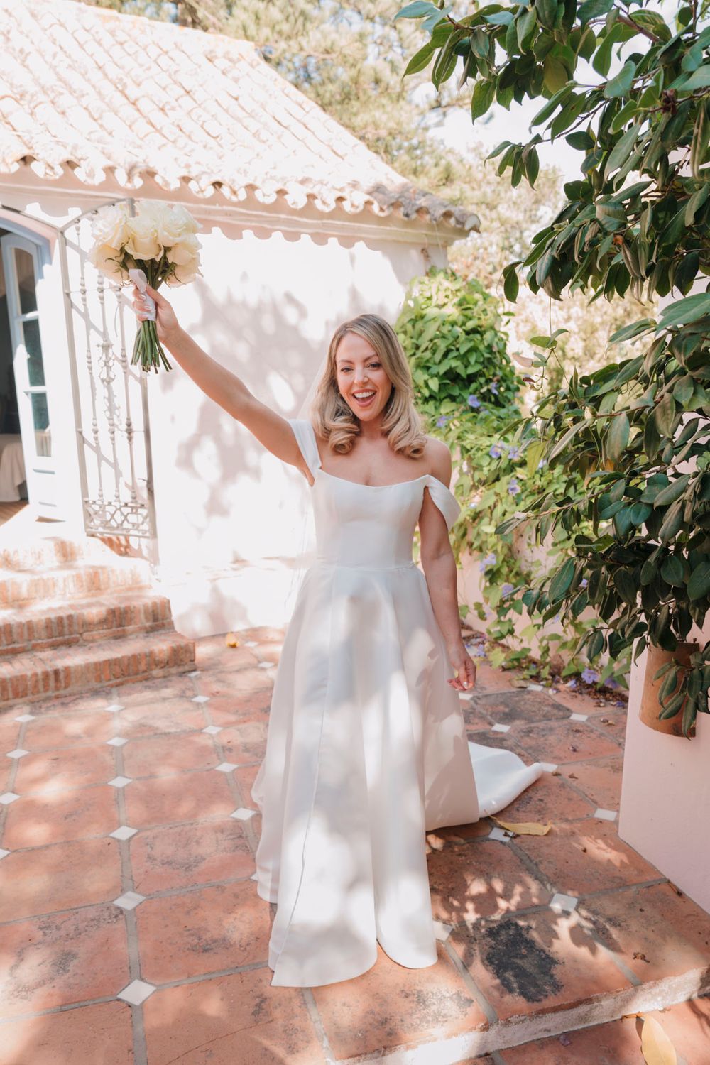 Joyful bride raises bouquet while standing on terracotta tiles in Mediterranean setting.