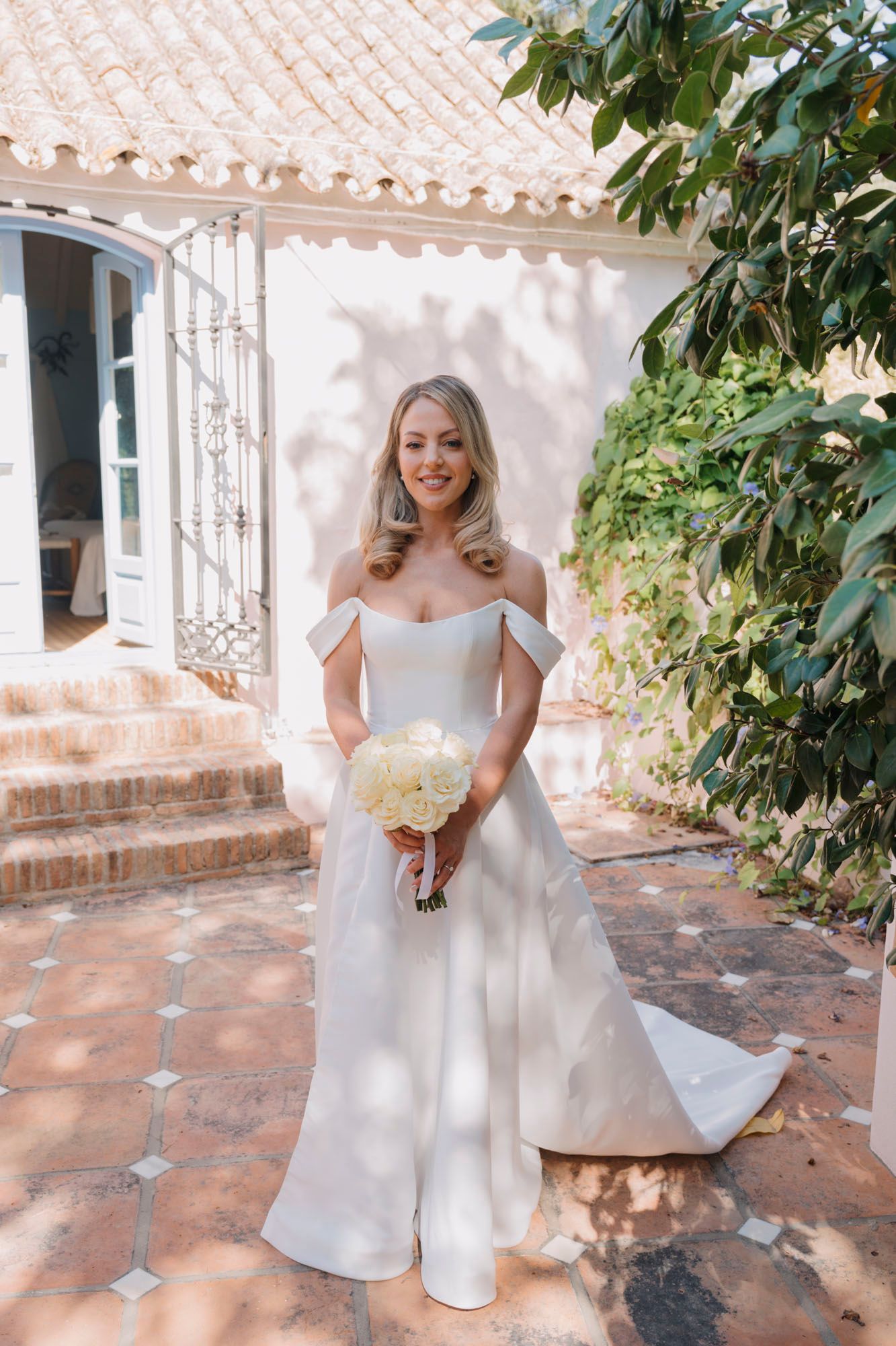 A bride in an elegant off-shoulder white wedding gown holds a cream bouquet on a terracotta patio.
