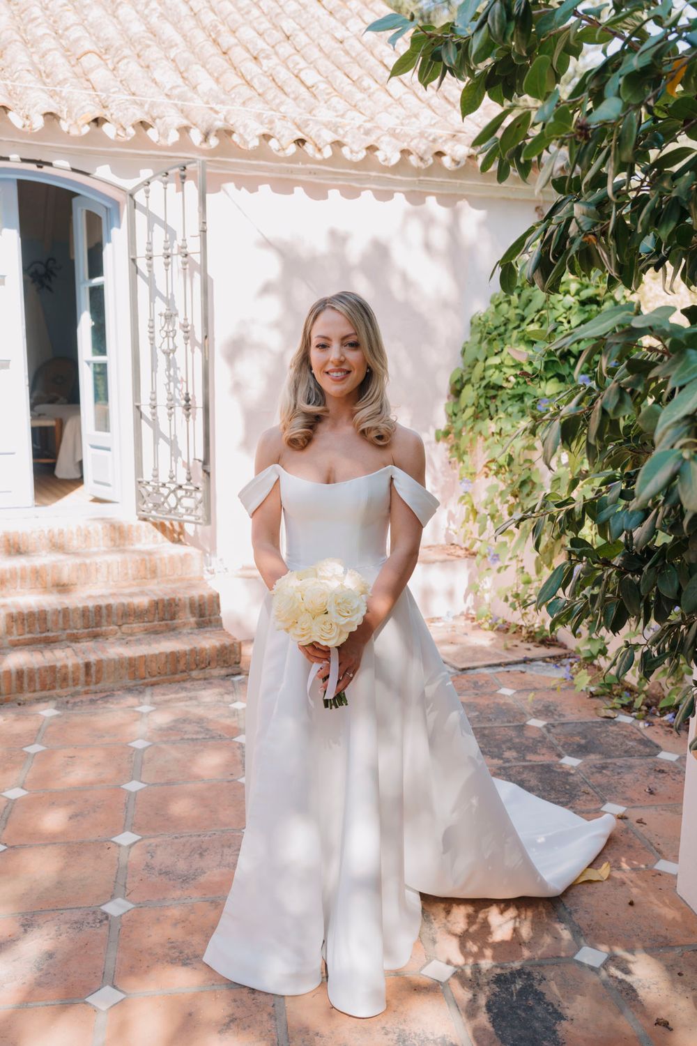 A bride in an elegant off-shoulder white wedding gown holds a cream bouquet on a terracotta patio.