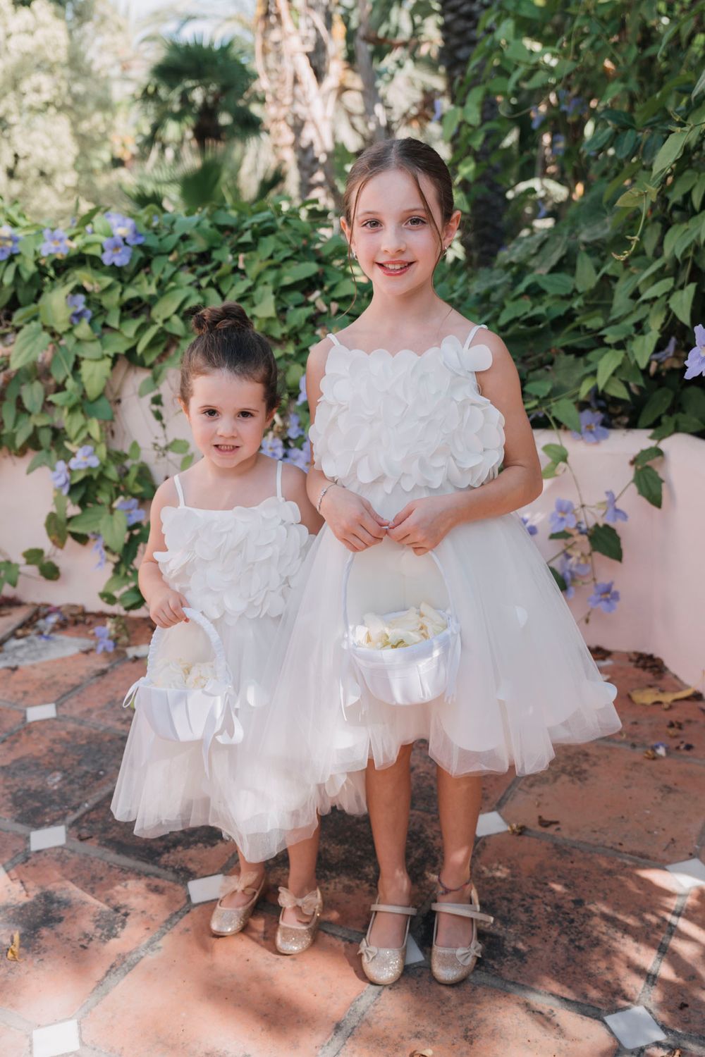 Two young flower girls in white tulle dresses pose on a brick patio with flower baskets.