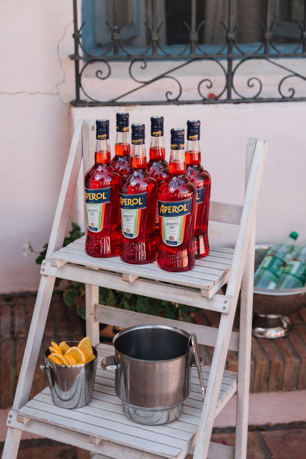 Red Aperol bottles displayed on white wooden ladder shelf with citrus and ice bucket.