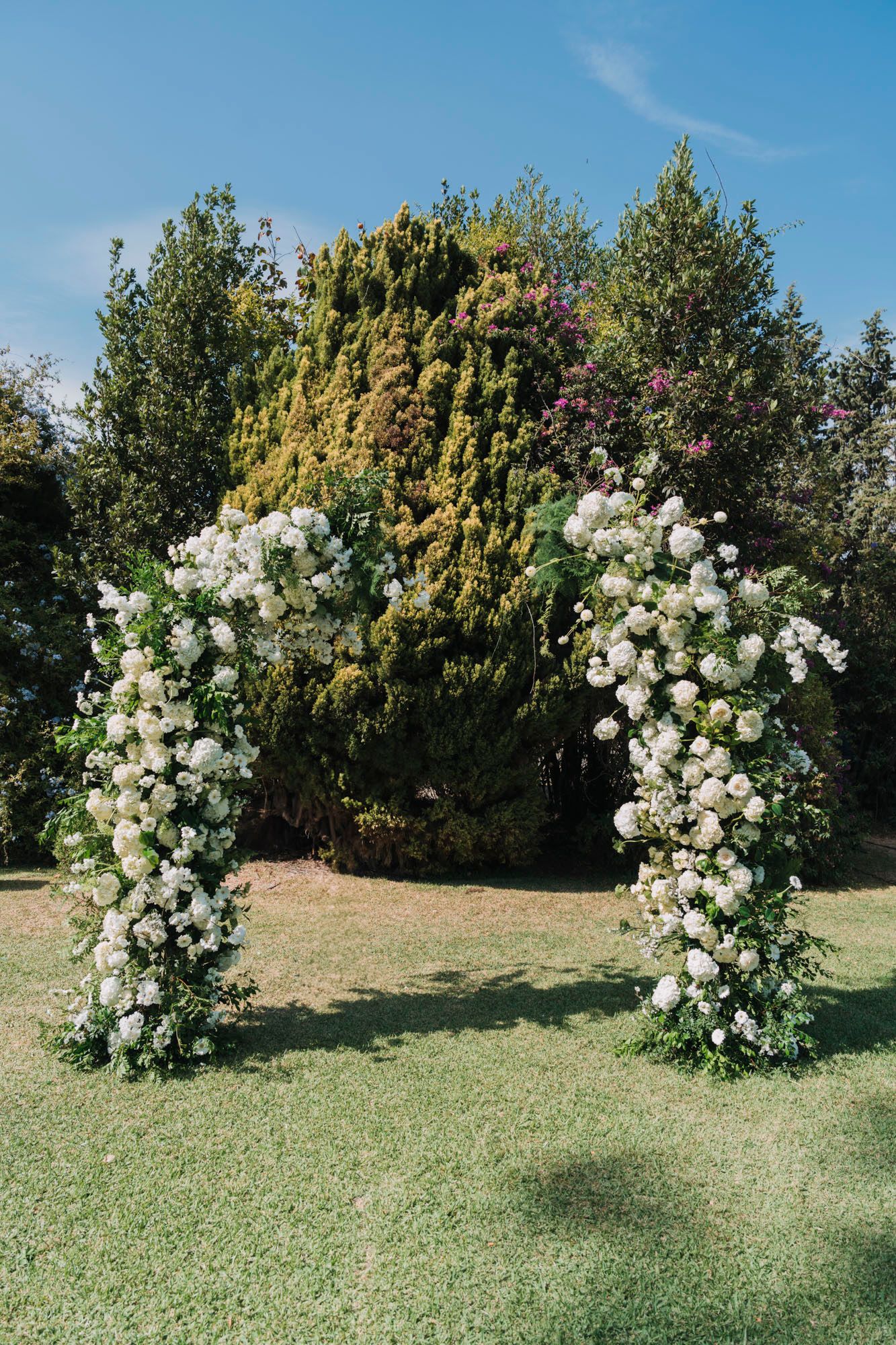 White floral arch made of roses stands on a green lawn for a wedding ceremony.