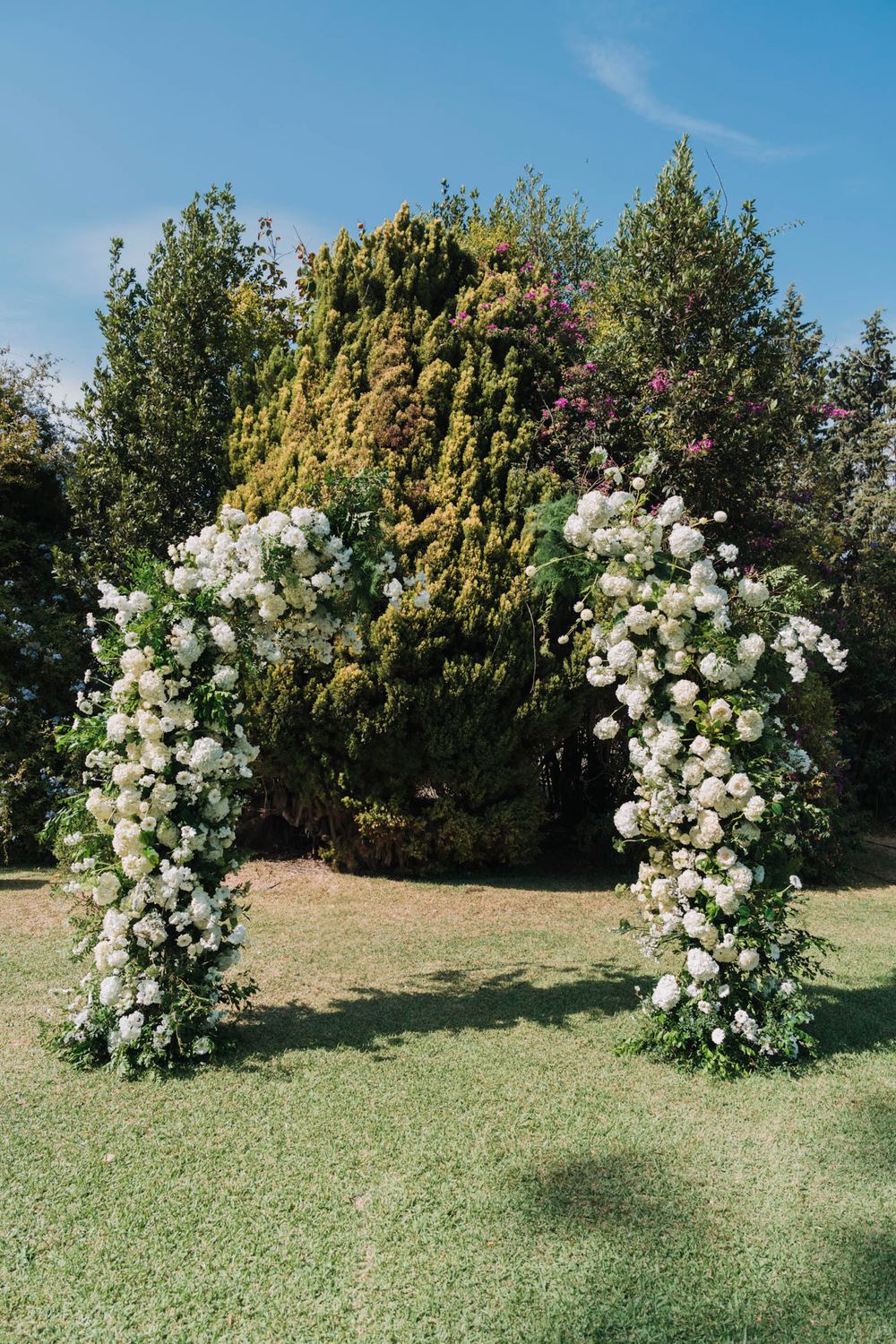 White floral arch made of roses stands on a green lawn for a wedding ceremony.