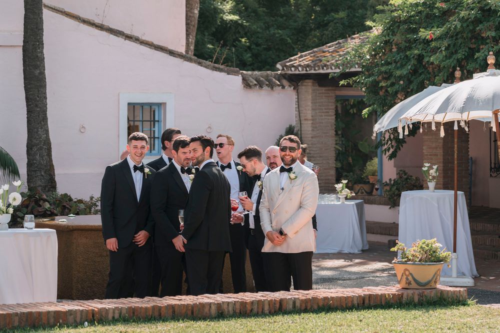Groomsmen in dark suits lined up at an outdoor wedding venue.