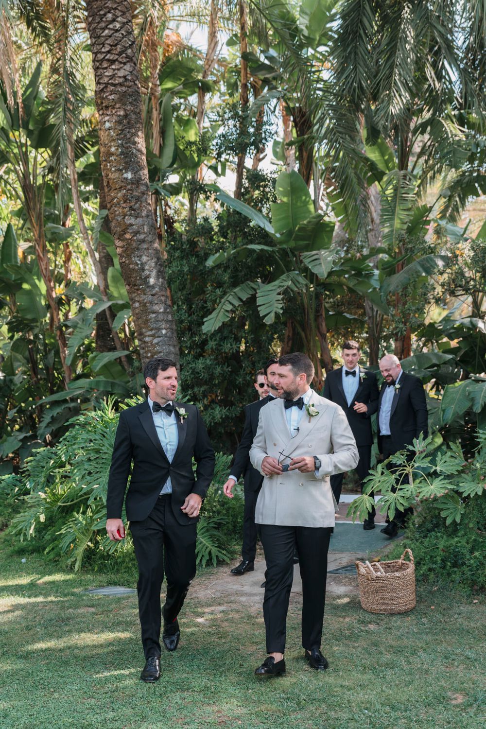Wedding party walking along a tropical garden path lined with palm trees and lush greenery.