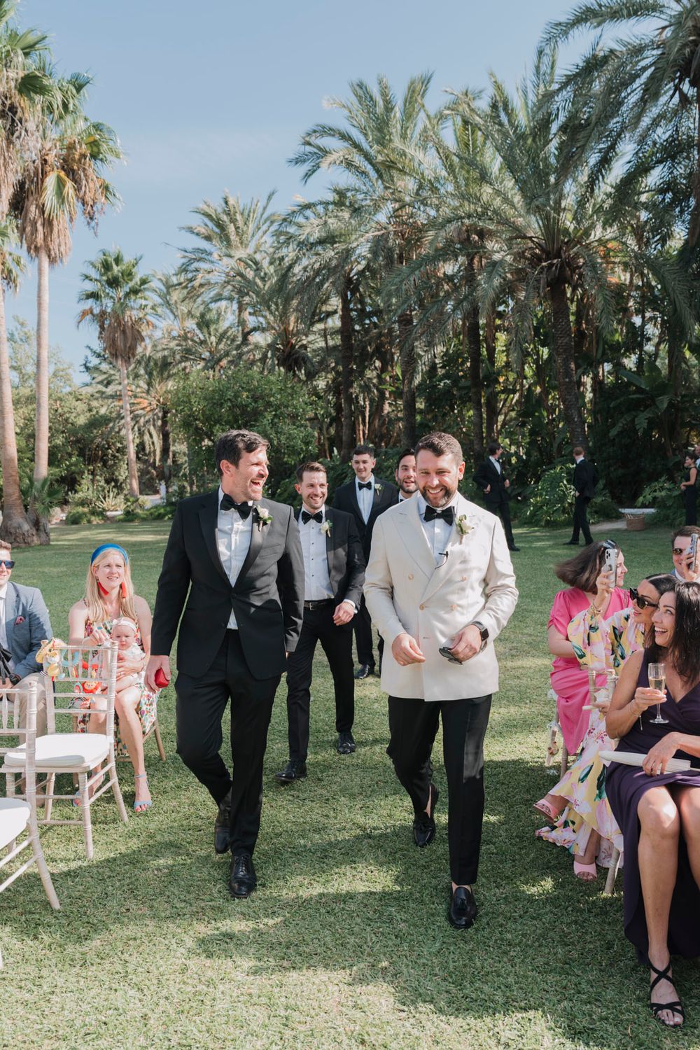 Elegant wedding ceremony processional through a palm tree garden with guests seated on both sides.