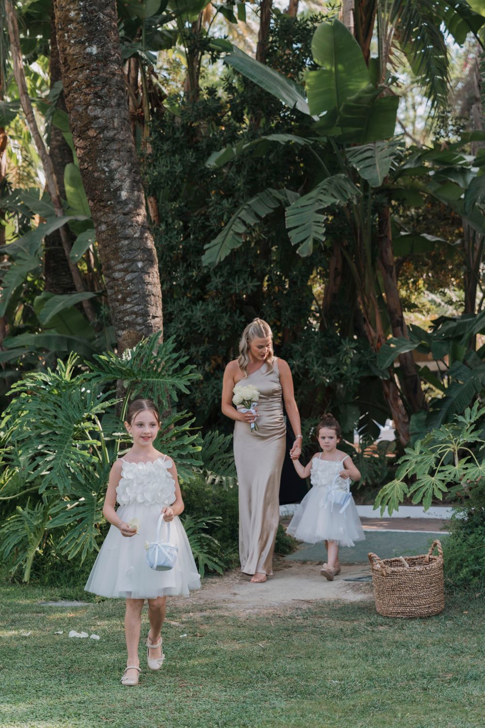 Flower girls in white dresses walking down a tropical garden path during a wedding ceremony.