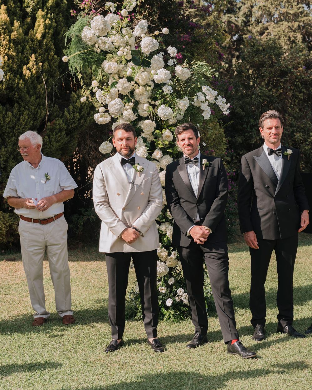 Wedding party members standing by a beautiful white floral arch in an outdoor garden setting.