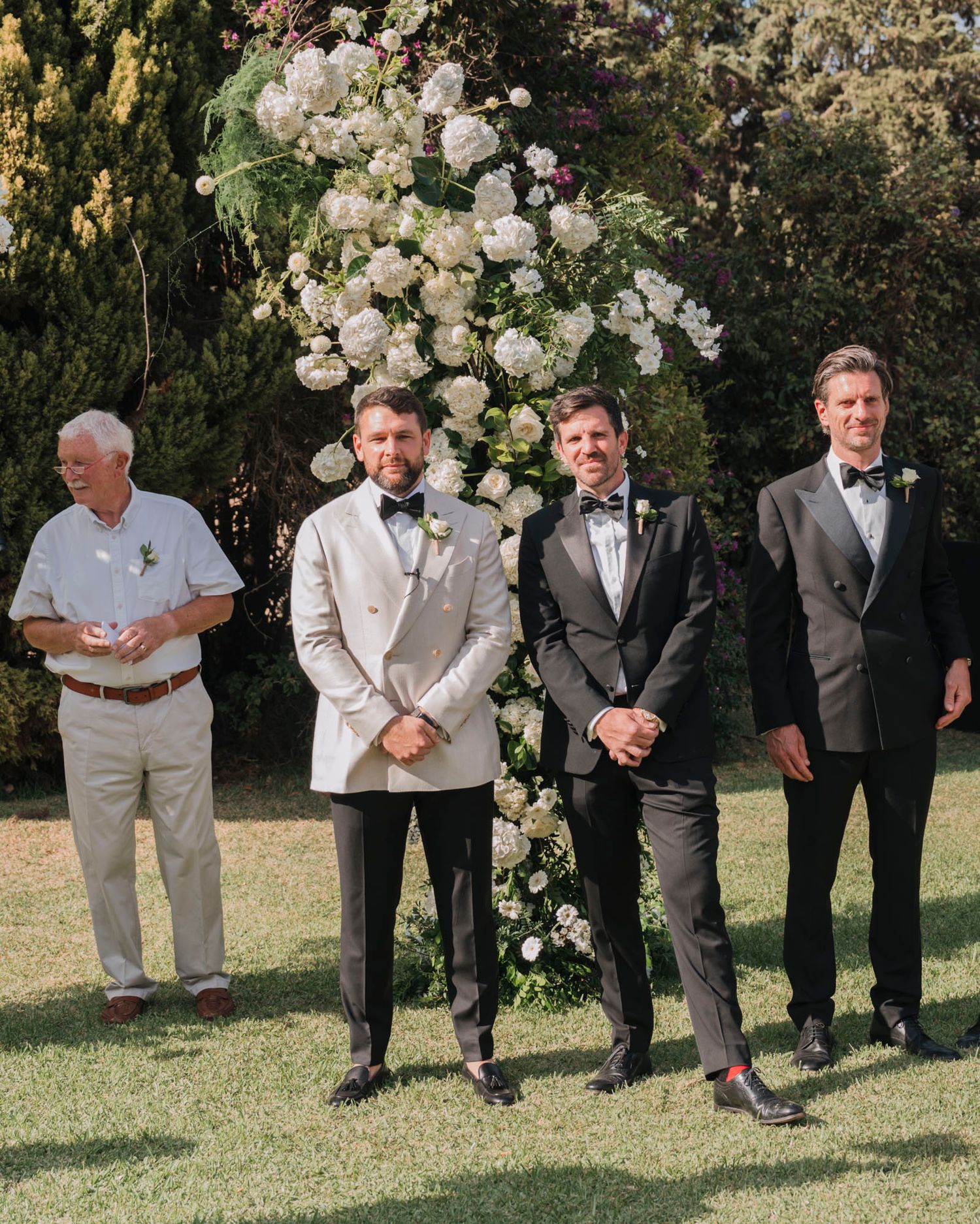 Wedding party members standing by a beautiful white floral arch in an outdoor garden setting.