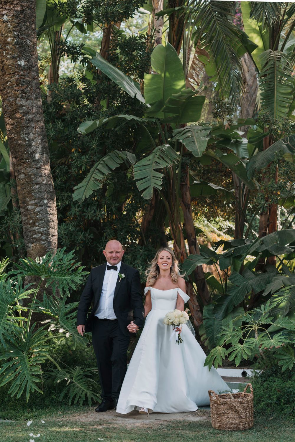 Wedding couple emerging from a lush tropical garden setting with palm trees and banana leaves.