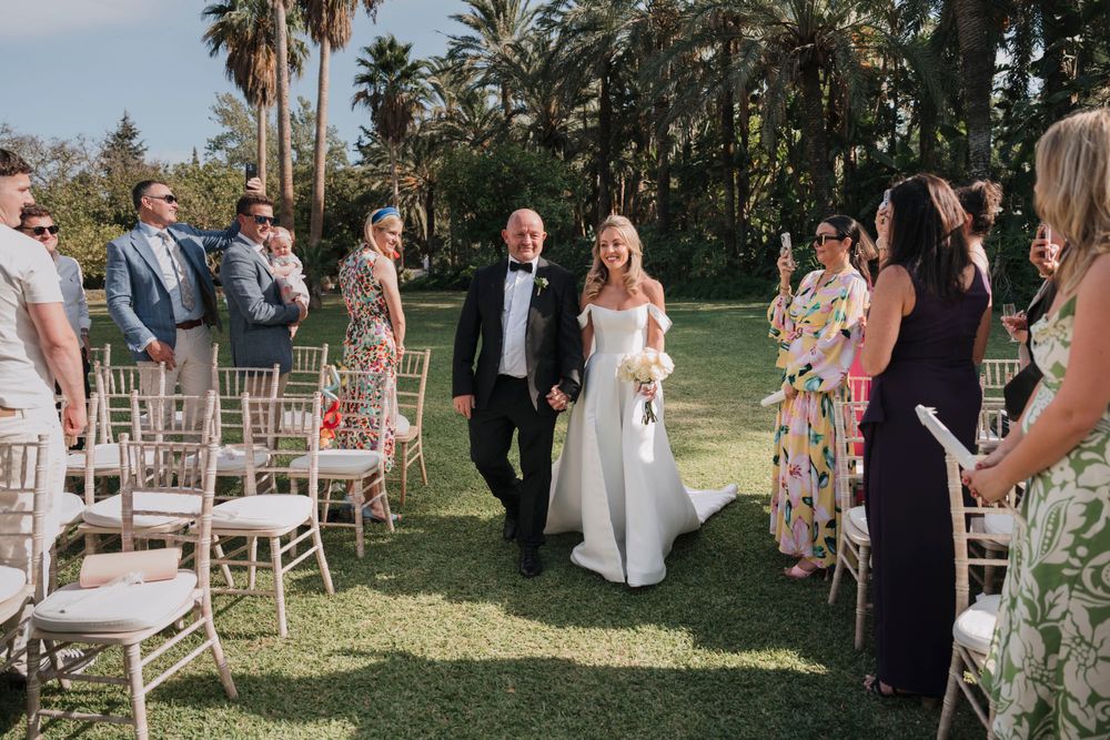 Wedding ceremony recessional with guests seated on white chairs in a tropical garden setting.