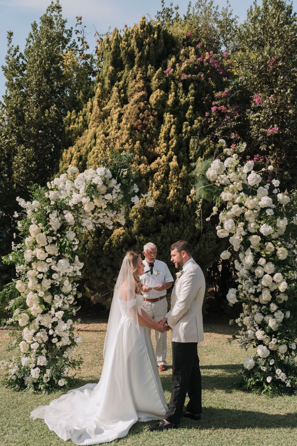 Wedding ceremony under an elegant white floral arch surrounded by tall trees and greenery.