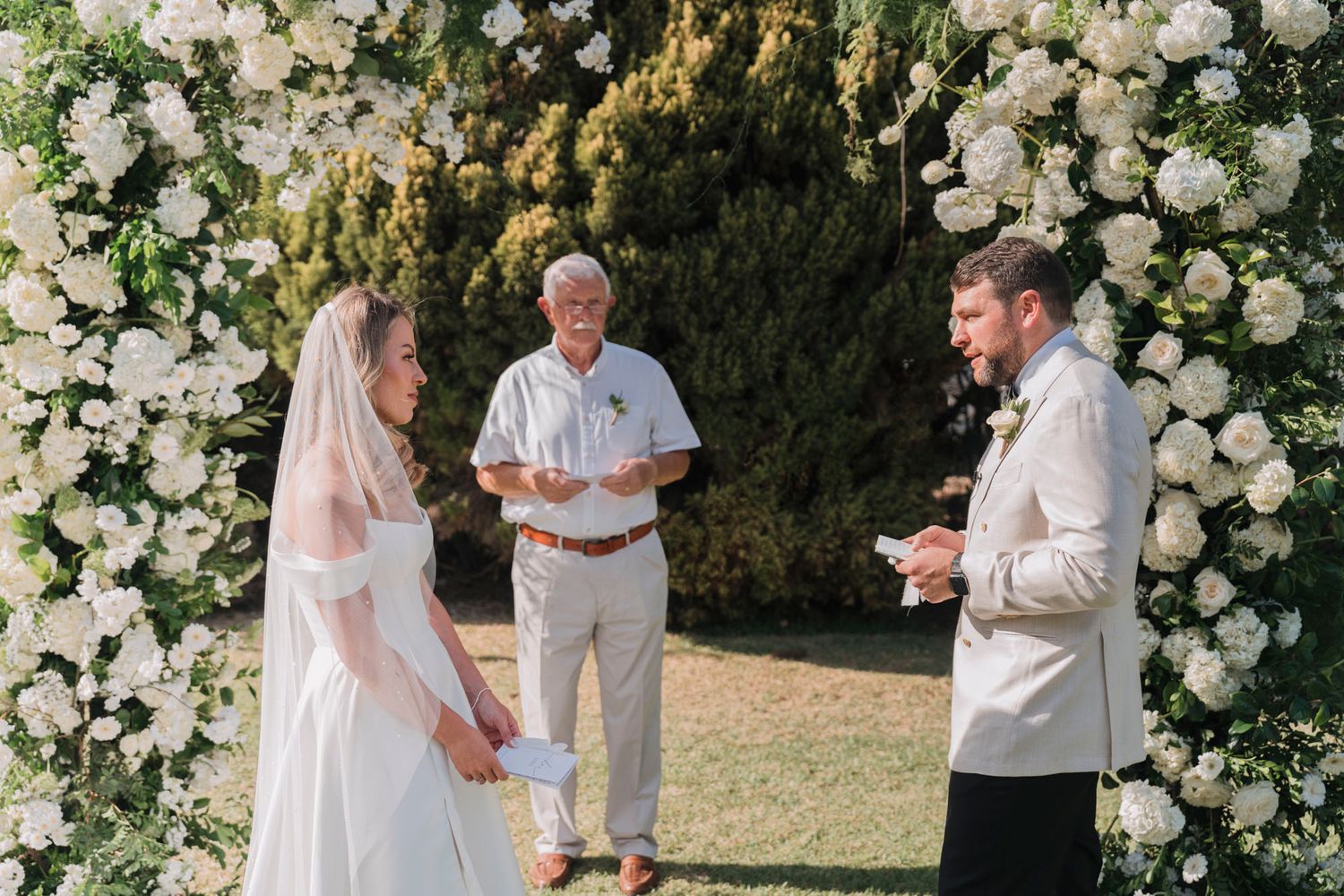Wedding ceremony taking place beneath a stunning white rose arch in a garden setting.