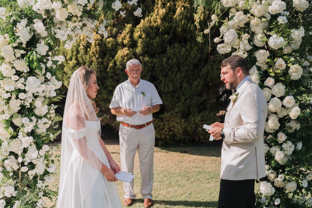 Wedding ceremony taking place beneath a stunning white rose arch in a garden setting.