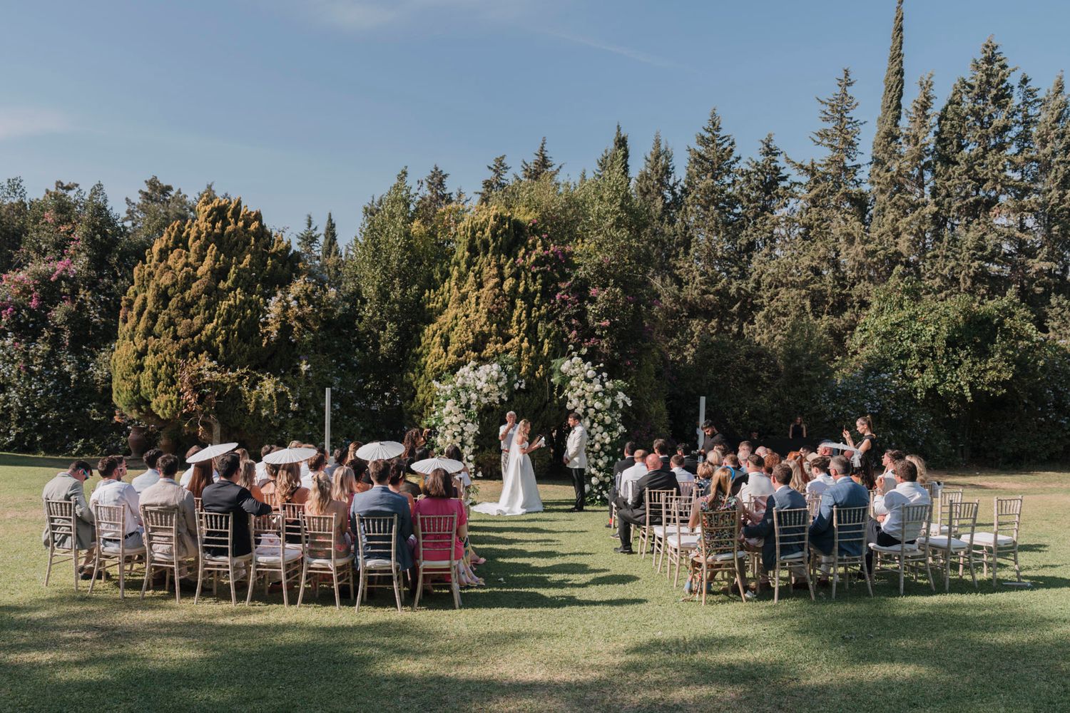 Scenic outdoor wedding ceremony with guests seated on white chairs surrounded by towering trees.