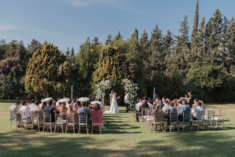 Scenic outdoor wedding ceremony with guests seated on white chairs surrounded by towering trees.
