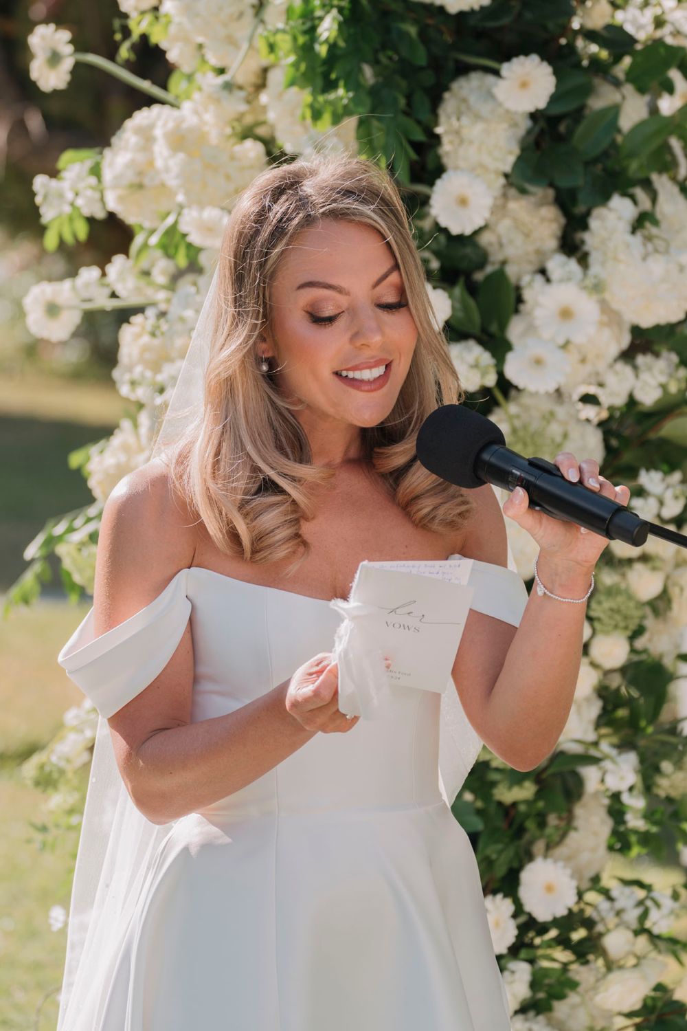 A woman in a white dress delivers a speech against a backdrop of white roses at an outdoor wedding ceremony.